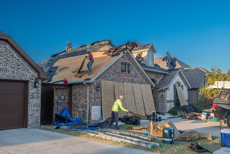 Roofing crew replacing the shingles on a brick house, with materials on the driveway